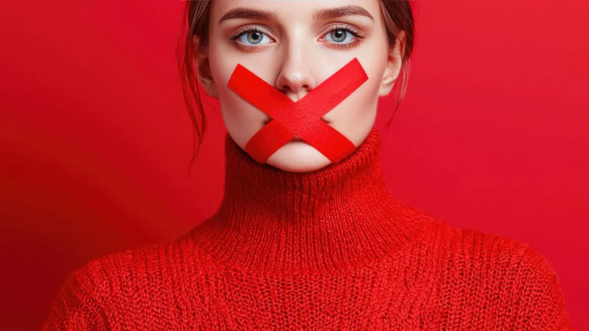 Kobieta w czerwonym swetrze na czerwonym tle z zaklejonymi ustami w kształcie krzyża, symbolizująca tłumienie emocji i brak wyrażania prawdy.